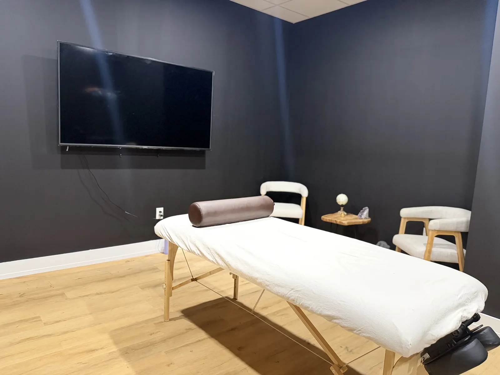Massage room with a made-up massage table, two chairs, a floor lamp and a wall-mounted TV